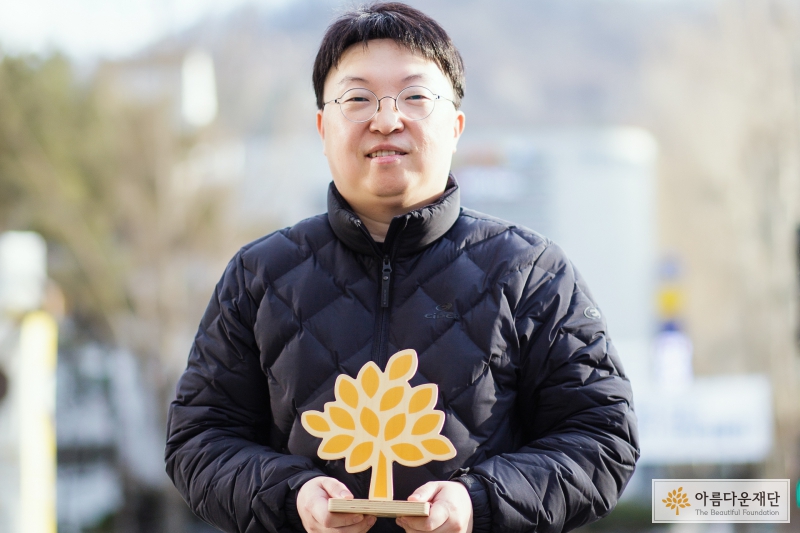 아름다운재단 씨앗나무(주황색으로 그려진 나무) 모형을 들고 길 가에 서 있는 정경찬 기부자. 밝게 웃고 있다. 