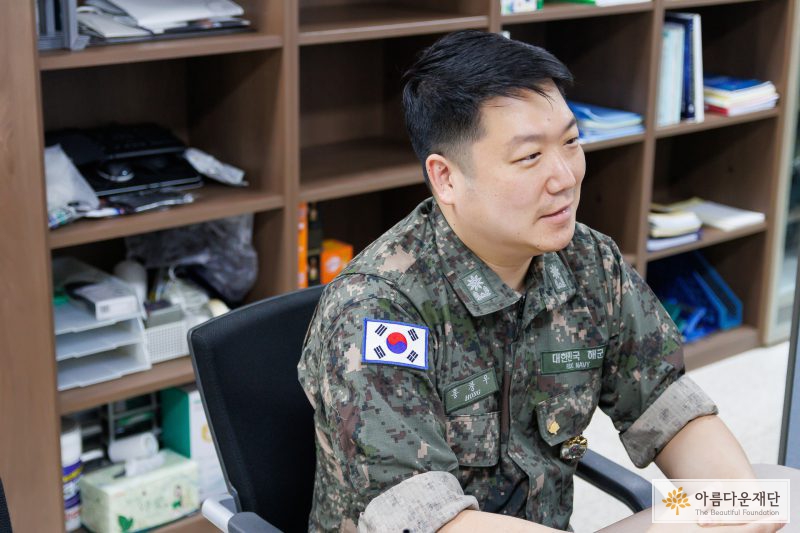 홍창우 기부자가 인터뷰를 하면서 밝게 웃고 있다. 양 손은 모으고 바른 자세로 의자에 앉아 있다. 기부자의 왼쪽 가슴에는 아름다운재단 6년 차 배지가 달려 있다. 