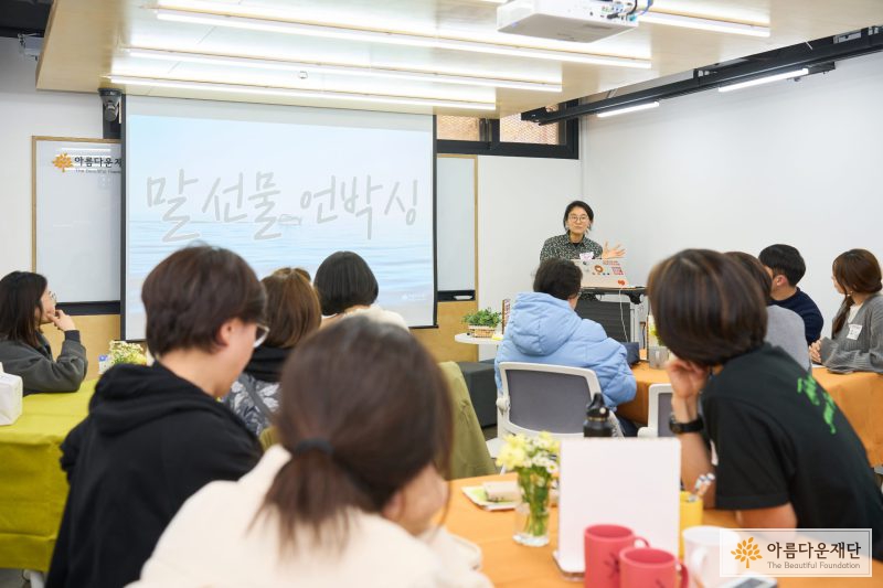 한 사람이 서서 발표하고 있고, 참가자들이 그를 보며 이야기를 듣고 있다. 