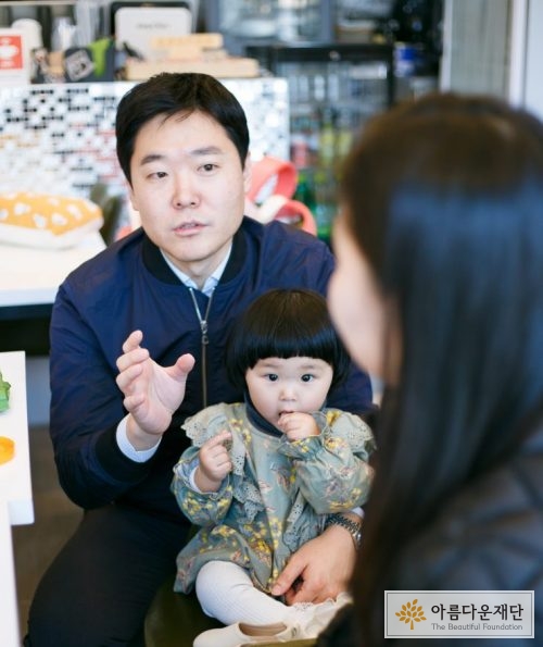 정면에 남색 자켓을 입은 남자가 반대편 여자를 바라보며 이야기 하고 있고, 남자 앞에 여자 아이가 입에 손을 물며 앉아 있다