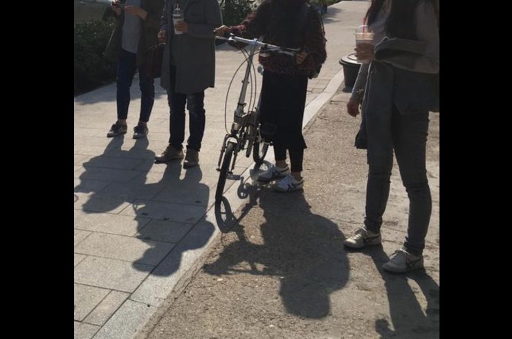 어깨 아래로 구도를 잡아 찍은 네 명의 사람이 있는 이미지. 두 명은 한 손에 음료를 들고 있고, 한 명은 자전거 핸들을 잡고 있다.