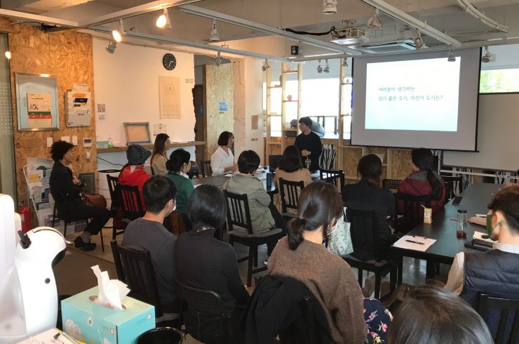 아늑해보이는 공간에 사람들이 앉아있고, 앞쪽에는 진행을 하는 듯 보이는 사람이 있다. 공간 앞 화면에는 '여러분이 생각하는 걷기 좋은 도시, 자전거 도시는?'이라고 적혀있다.